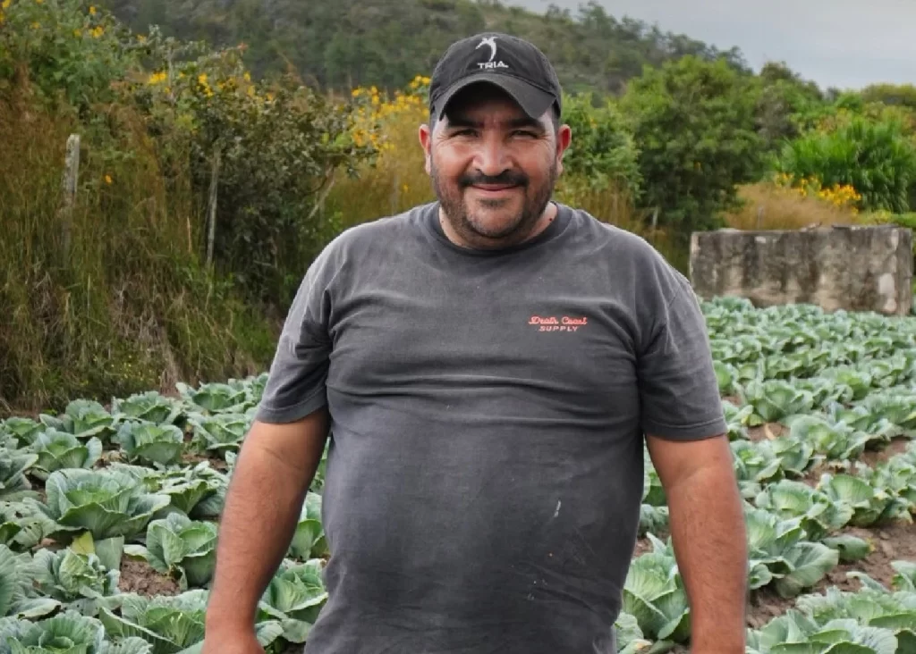 Un hombre con una camisa oscura y gorra está de pie, sonriendo en un campo de plantas verdes y frondosas, con arbustos y árboles al fondo.