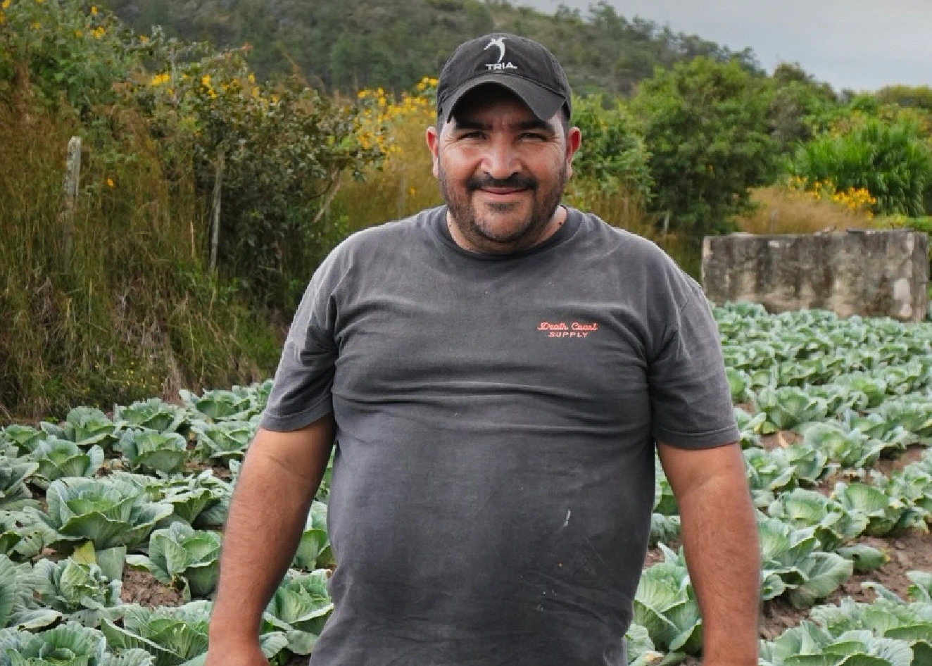 Un hombre con una camisa oscura y gorra está de pie, sonriendo en un campo de plantas verdes y frondosas, con arbustos y árboles al fondo.